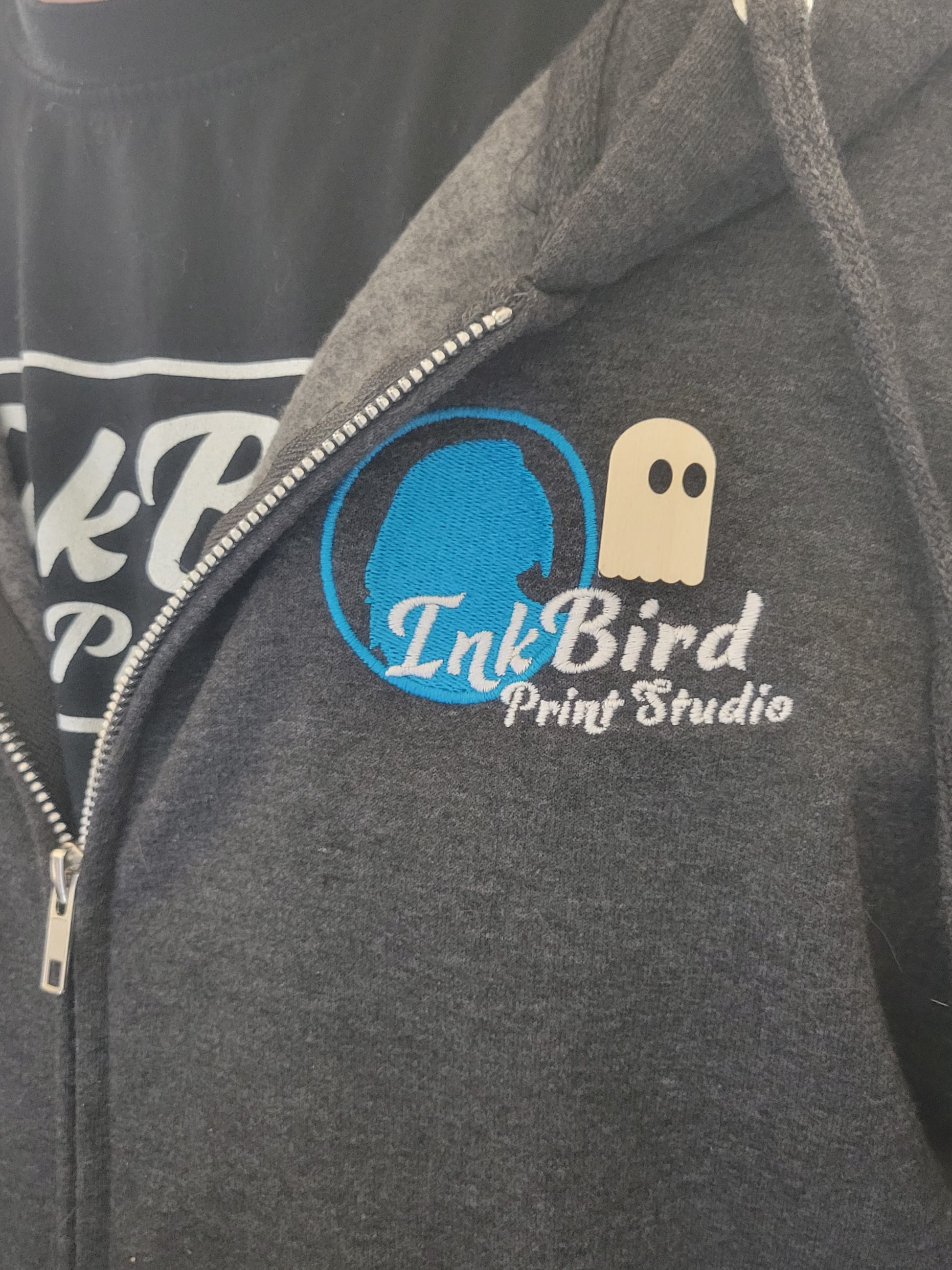 Dark gray zip-up hoodie with blue and white InkBird Print Studio logo, birch wood Simple Ghost Pin above logo, worn over black shirt.