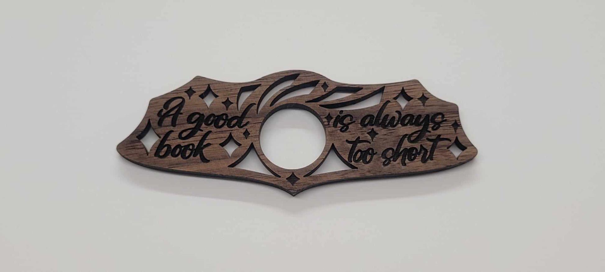 The Laser Cut Wood Book Holder from InkBird Print Studio LLC is crafted from walnut wood into intricate patterns, featuring the handwritten quote, A good book is always too short. Its an ideal thoughtful gift for any book lover.