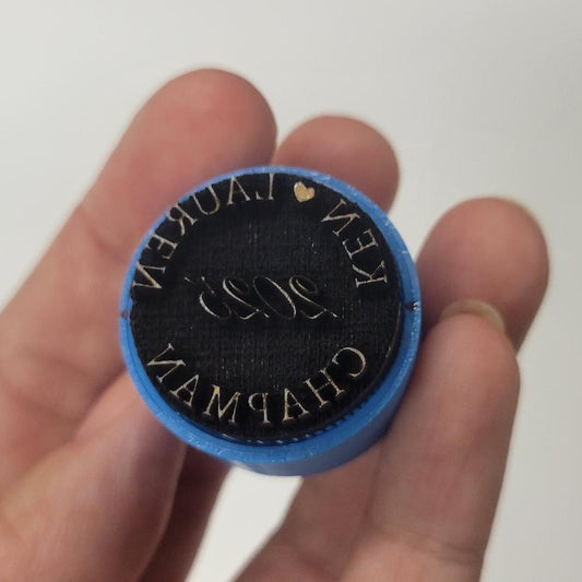 A hand holds a 3 round black ceramic makers mark stamp with blue edges, “KEN LAUREN CHAPMAN 2023” and heart engraving.